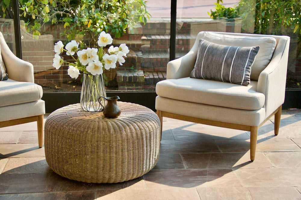 Pale upholstered chairs and a woven table beside sunroom windows, with white flowers on the table.