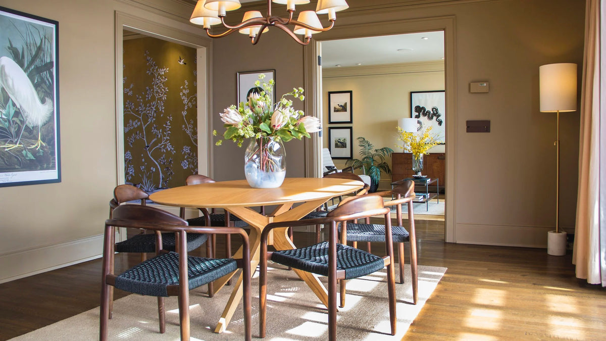 Wood dining chairs, a sculptural table and flowering branches in a sunlit room with traditional woodwork.