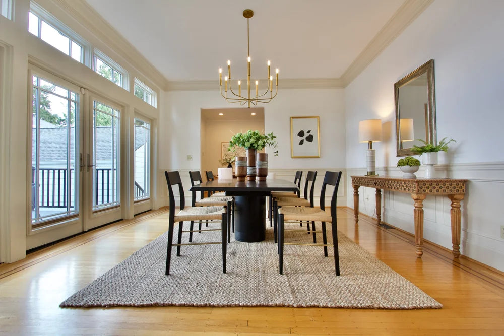 Woven dining chairs around a dark table, with brass lighting, a carved console and tall French doors.